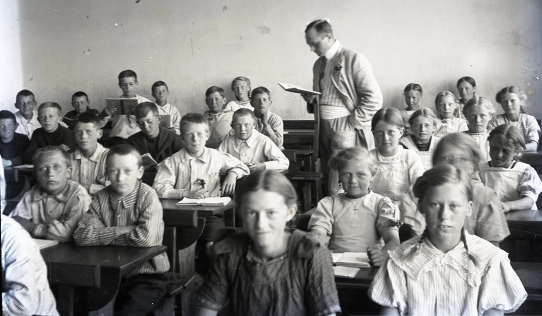 Historisk klassefoto: en skoleklasse ved deres pulte med læreren stående bagest med en bog i hånden.